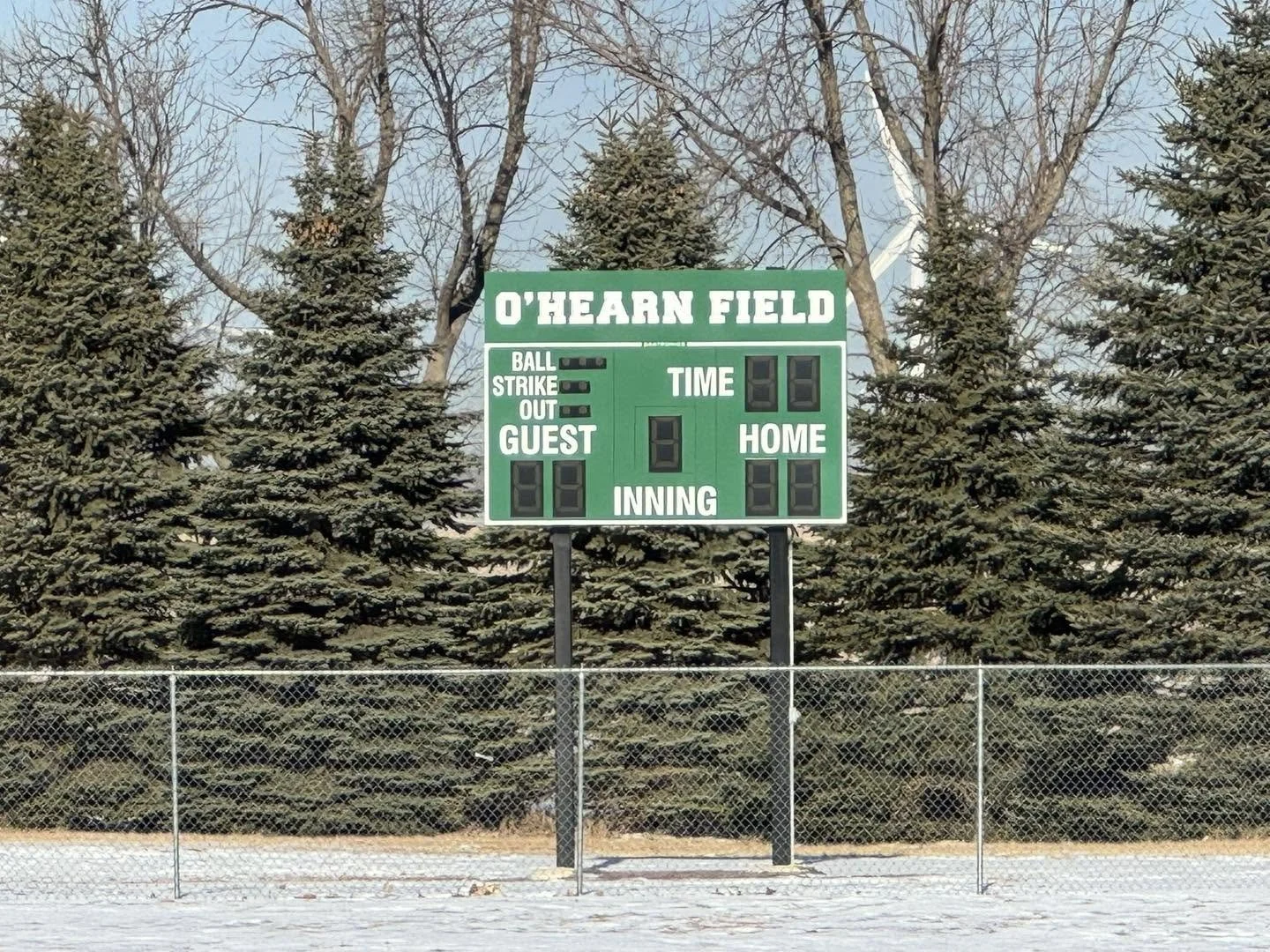 Green baseball scoreboard at O'Hearn Field with no game in progress, displaying 1 ball, 0 strikes, 1 out, 9 innings, 0 guest runs, 1 home run, 8 minutes and 1 second on the clock, and no score for away or home teams, surrounded by evergreen trees and a chain-link fence.