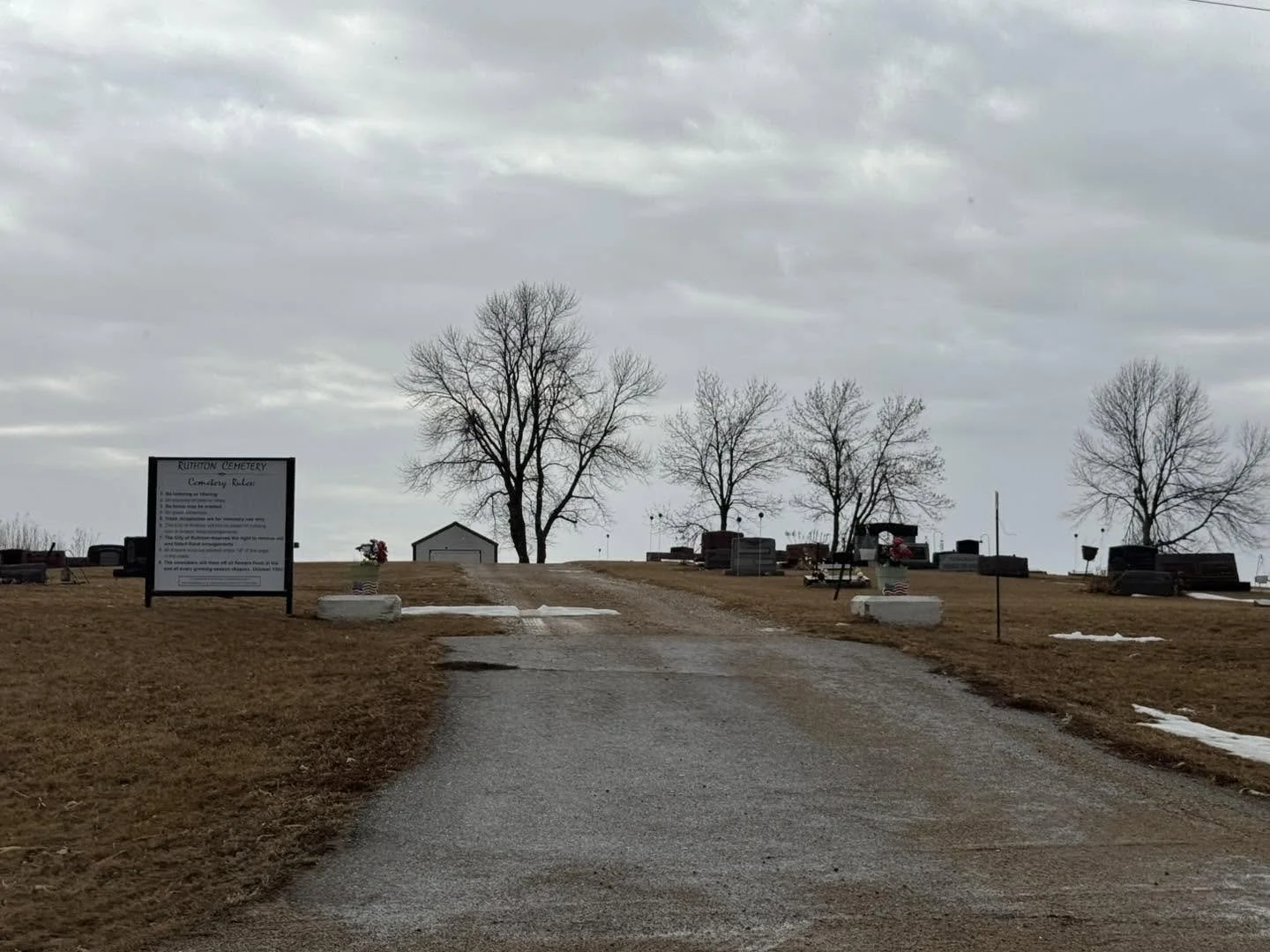 Gravel path leading to a cemetery with tombstones and leafless trees under an overcast sky.