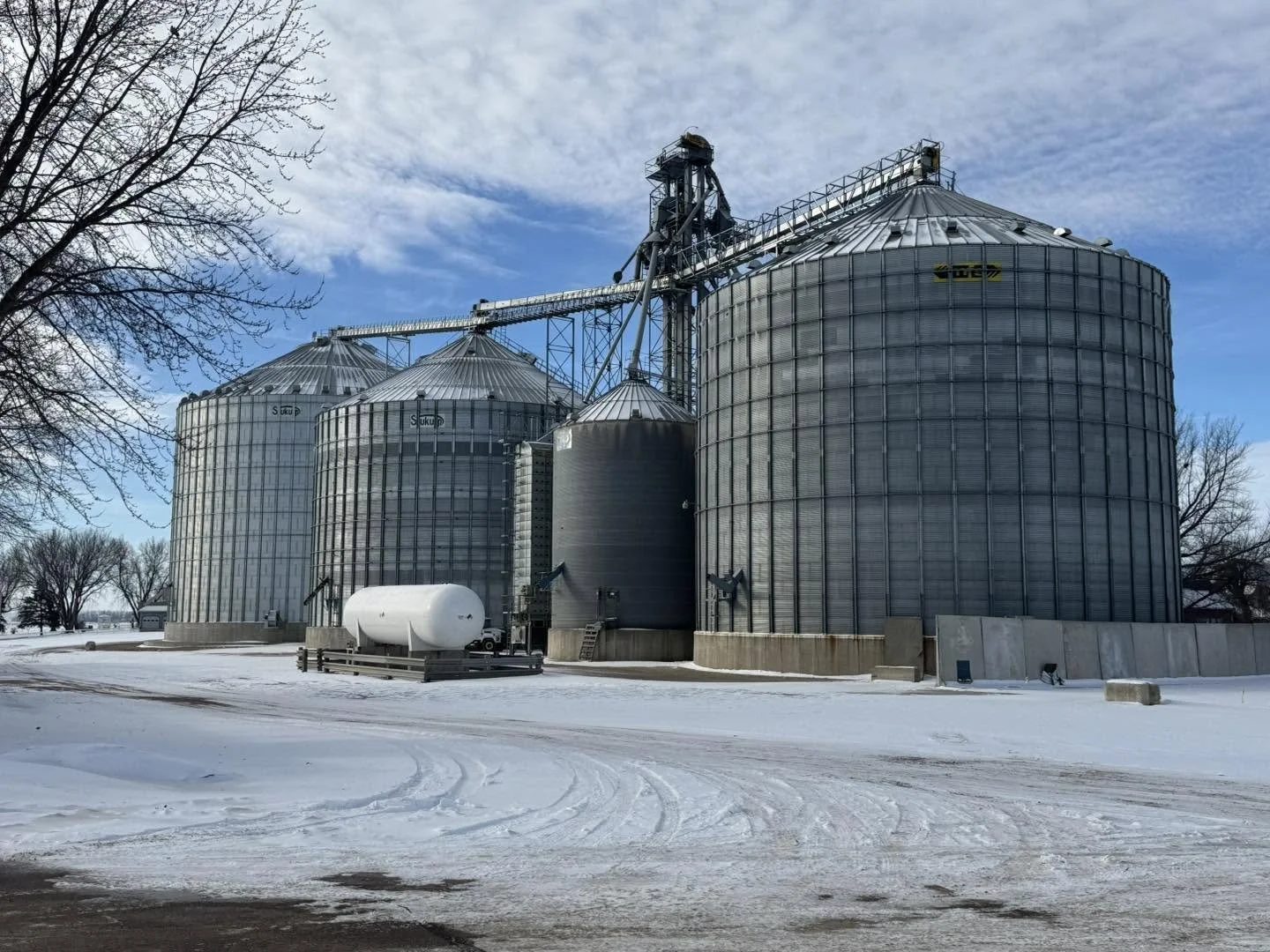 Four large metallic grain silos on a snowy farm under a partly cloudy sky with leafless trees in the background.