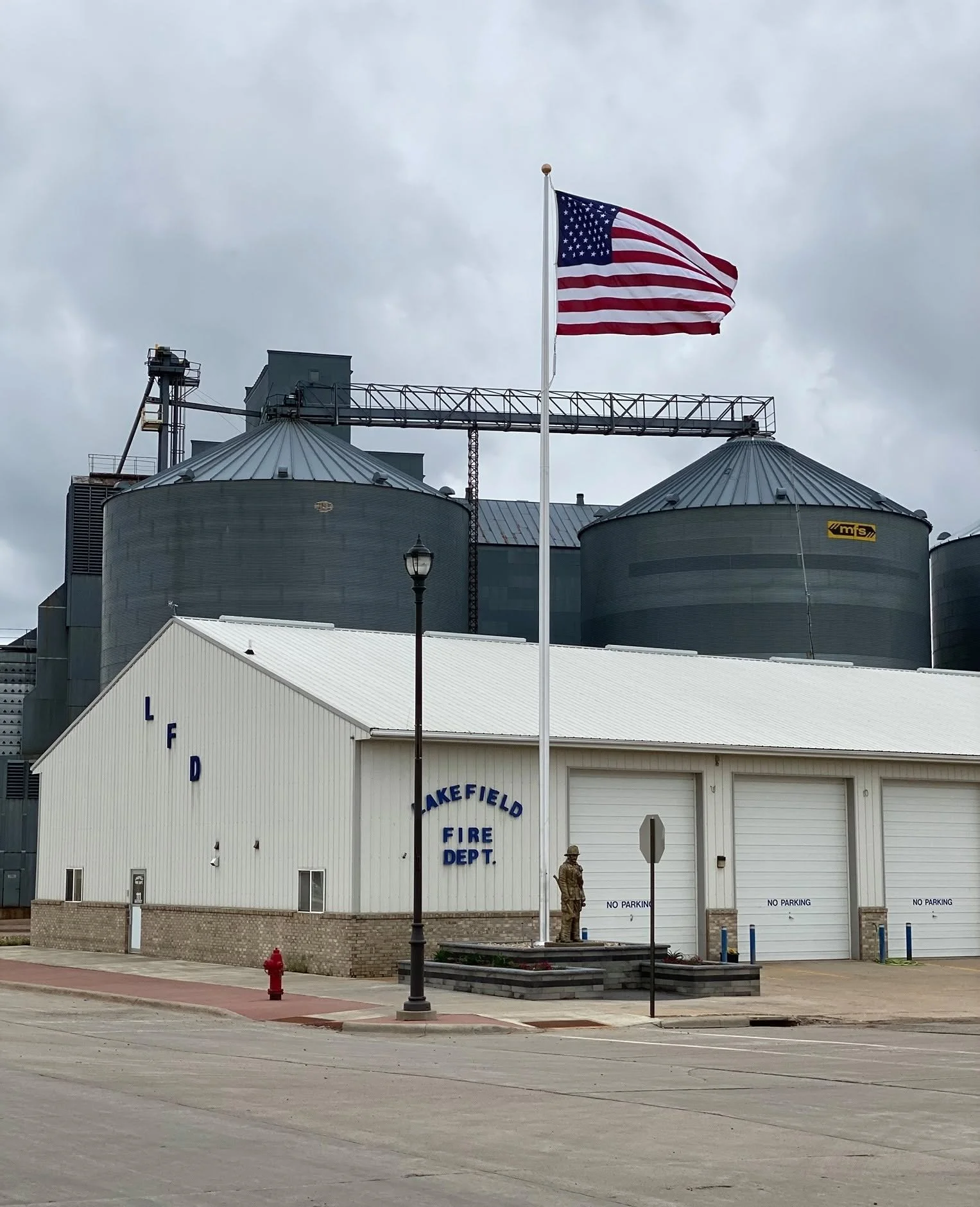 A white fire department building with a sign that reads "Lakefield Fire Dept." and a large American flag flying on a pole in front. There are two large storage silos behind the building, and a soldier standing near the entrance. There is a lamp post, a fire hydrant, and a curb with a sidewalk in the foreground.