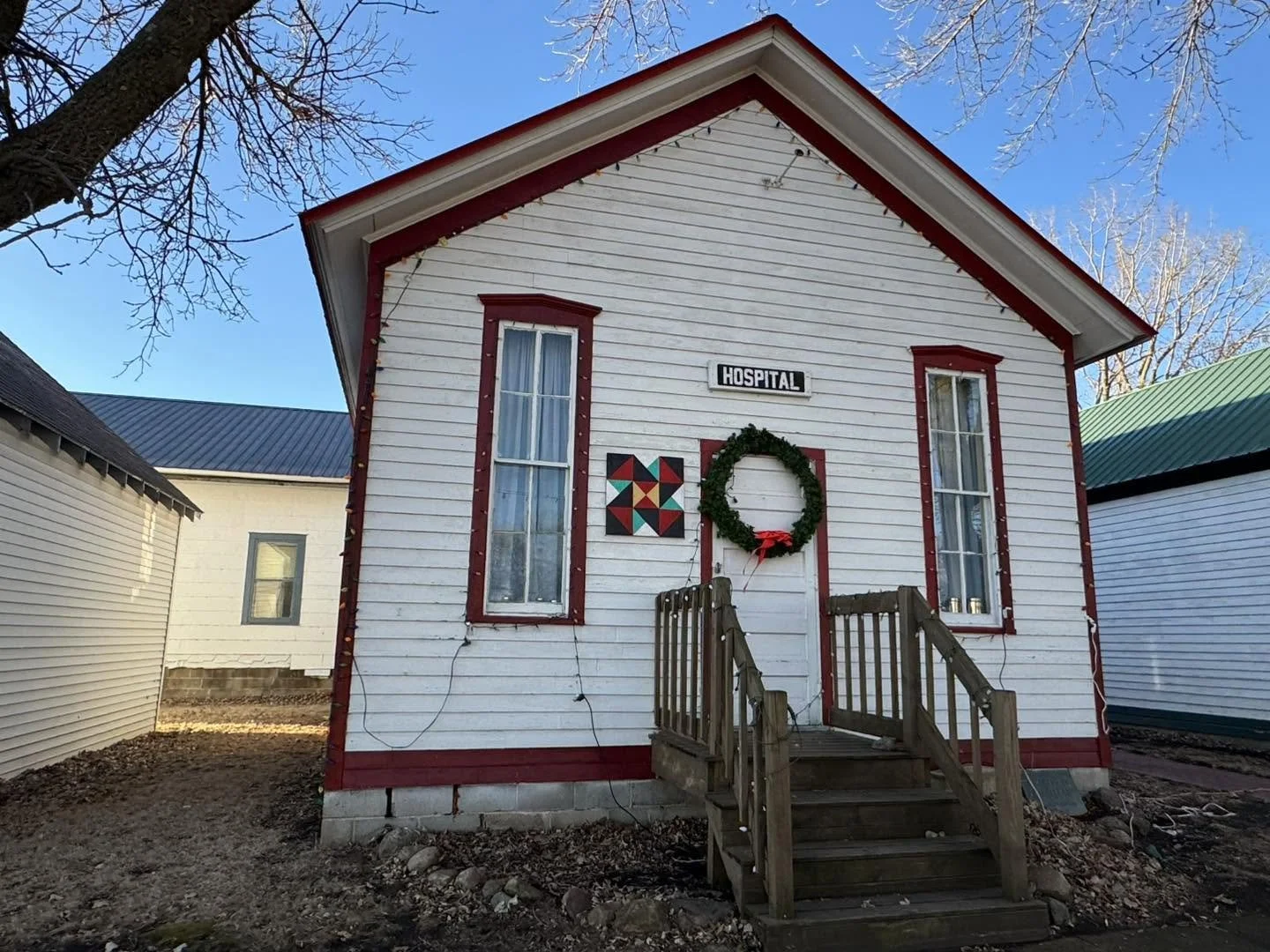 A small white wooden building decorated for Christmas, labeled as a hospital, with a wreath, string lights, and holiday decorations.