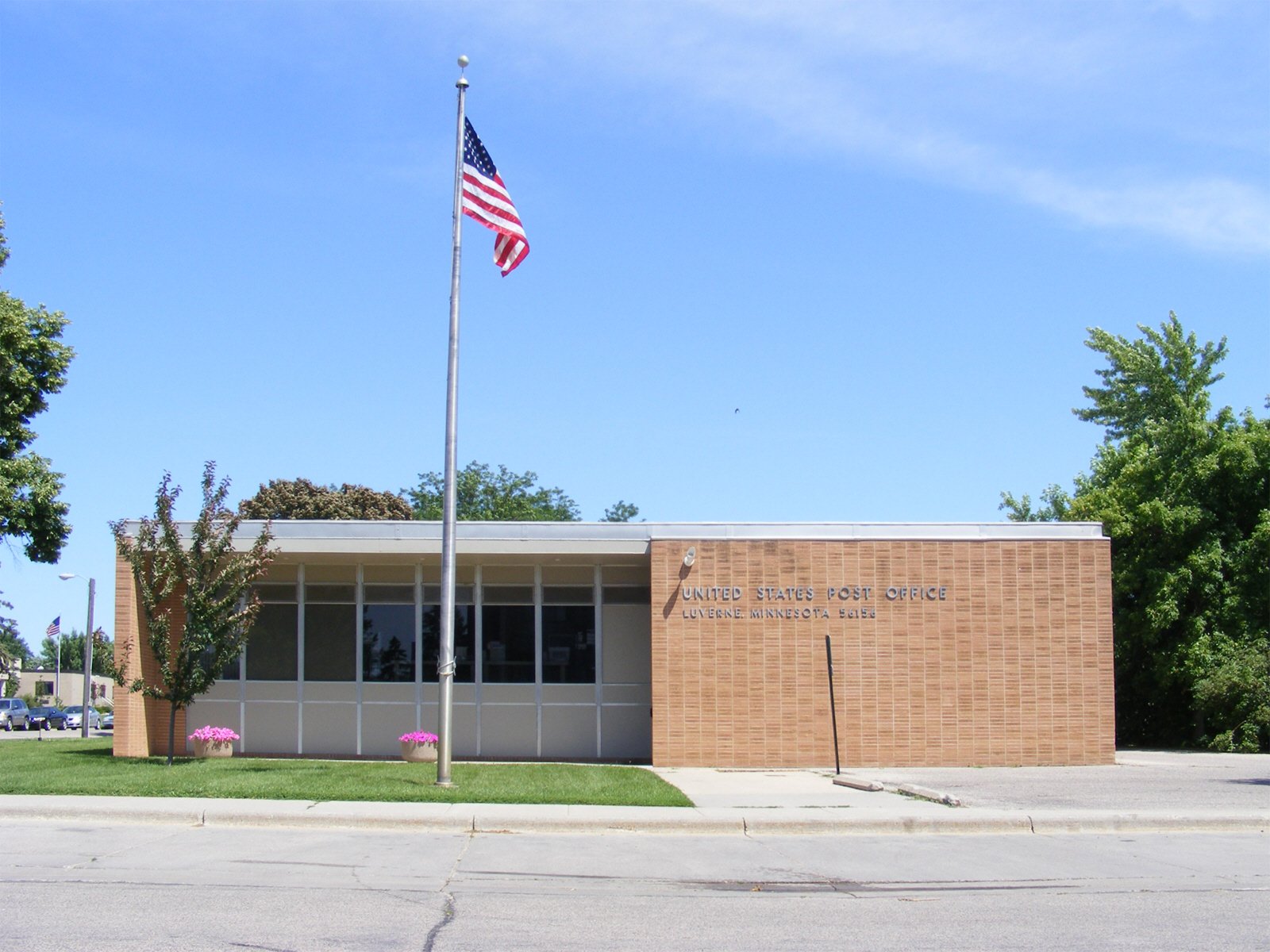 A small brick building labeled 'United States Post Office, Lure Minnesota 56284' with a flagpole flying the American flag in front of it. There are a few trees and pink flowers nearby and a parking lot to the side.