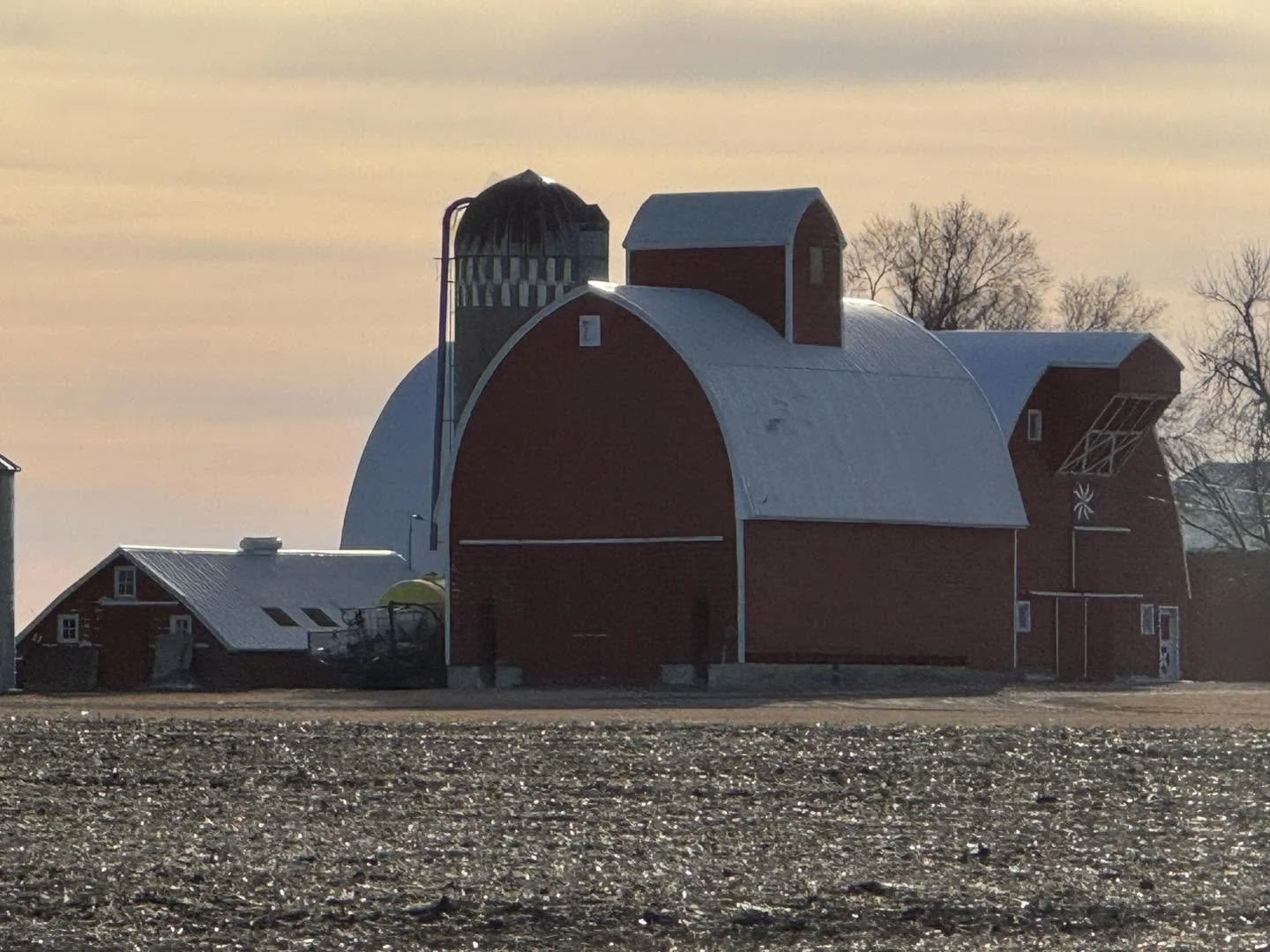 A large red barn with white accents and a curved roof, surrounded by leafless trees, under a sunset sky, with a gravel ground in the foreground.