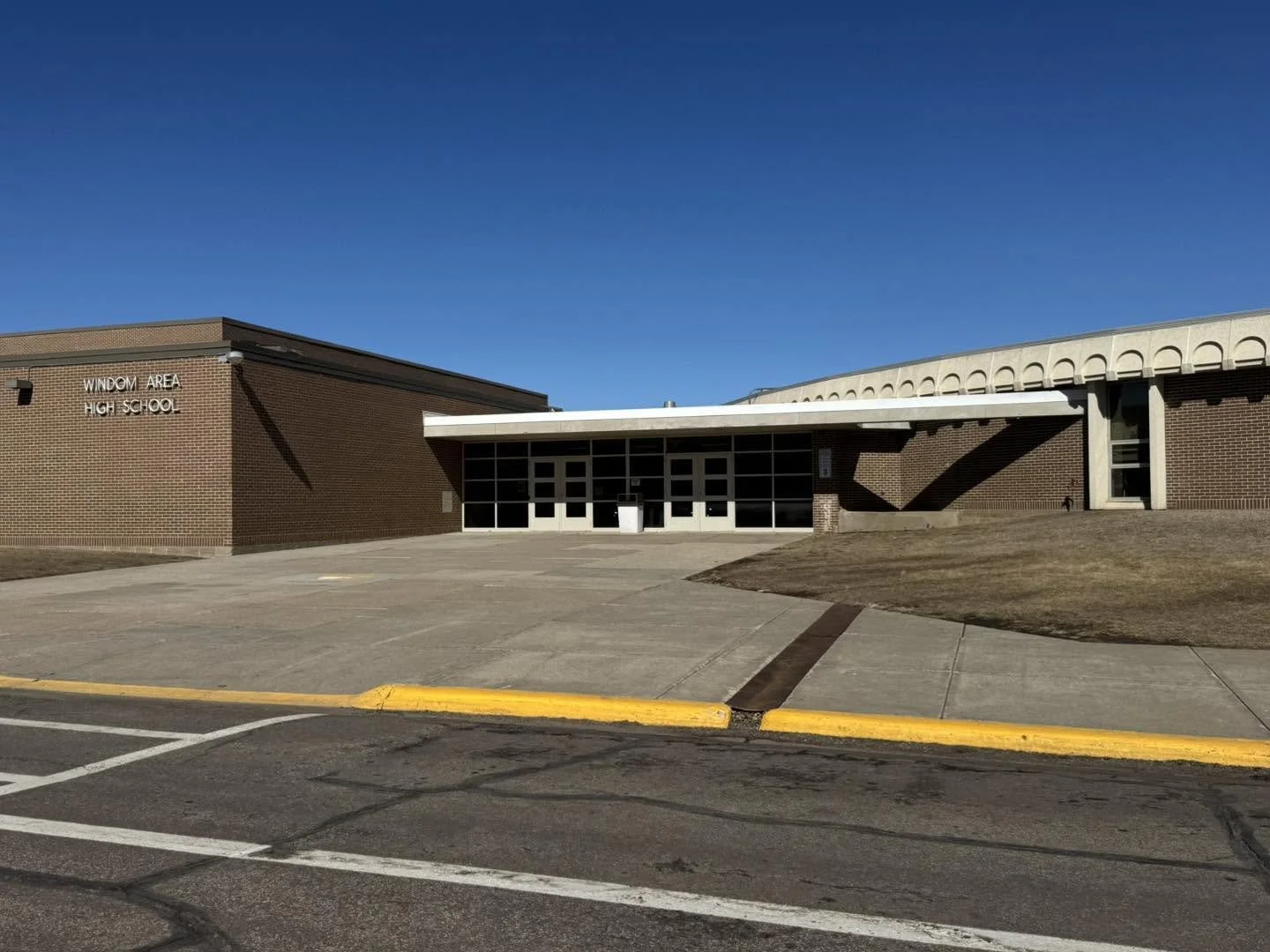 Front view of Windom Area High School building with brick exterior and glass entrance, under a clear blue sky, with empty parking lot in foreground.