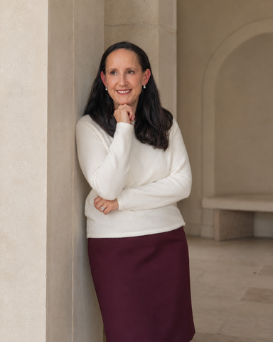 Ginny wears a white sweater and maroon skirt leaning against a wall in an indoor setting.