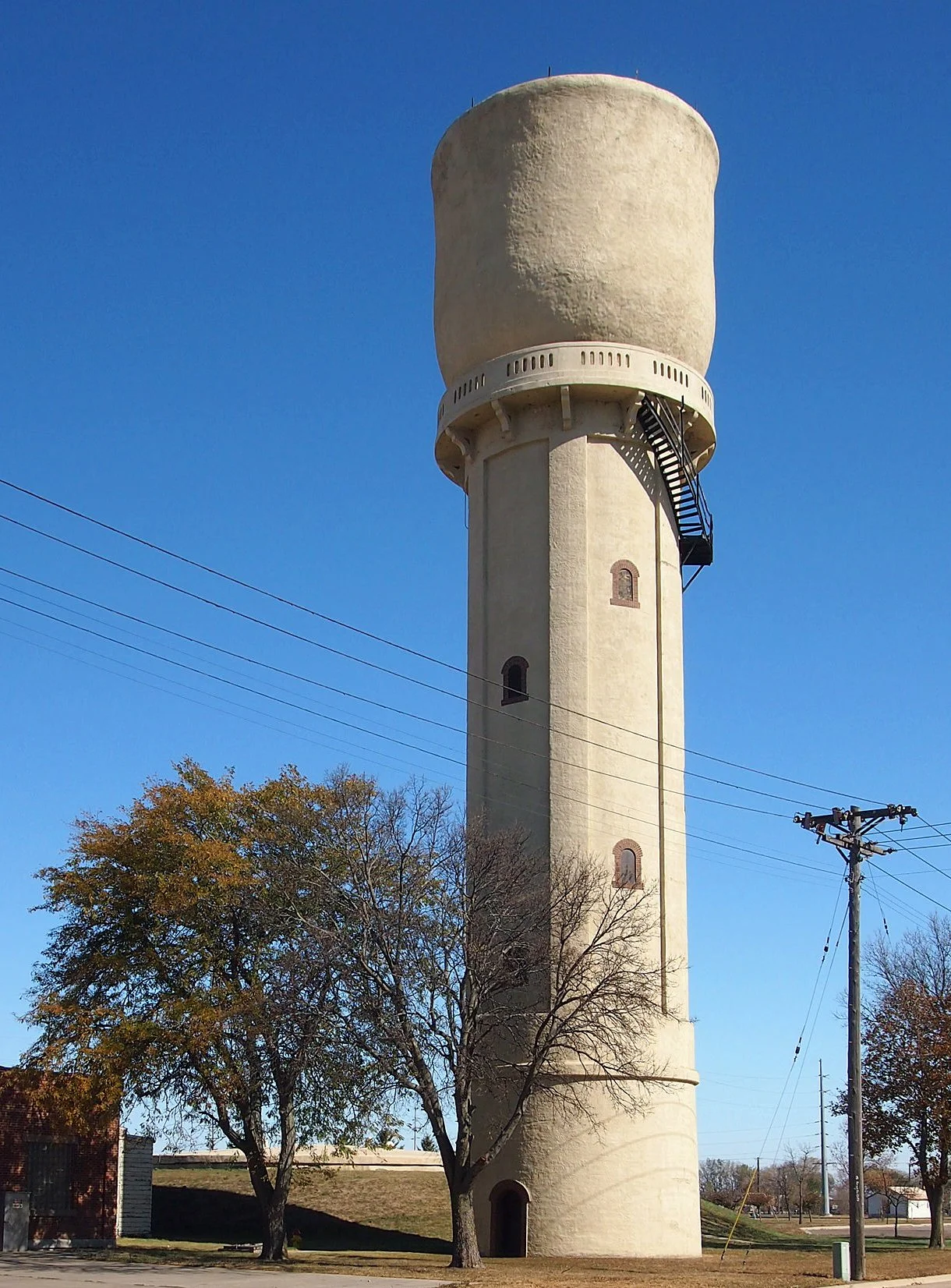A tall, beige water tower with a rounded top, located against a bright blue sky. There are trees with autumn foliage and power lines nearby.