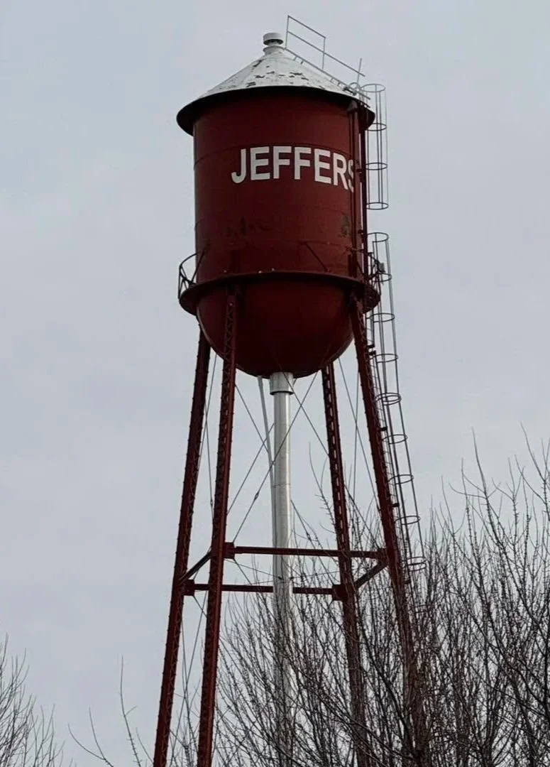 Red water tower with white letters spelling "JEFFER" on it, set against a cloudy sky. Bare tree branches are visible in the foreground.