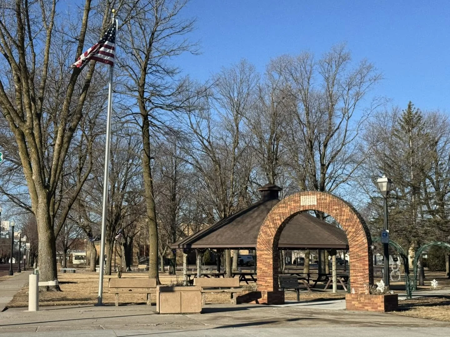 A park with leafless trees, a small brick and wood pavilion, a flagpole with an American flag, benches, and a lamp post, under a clear blue sky.
