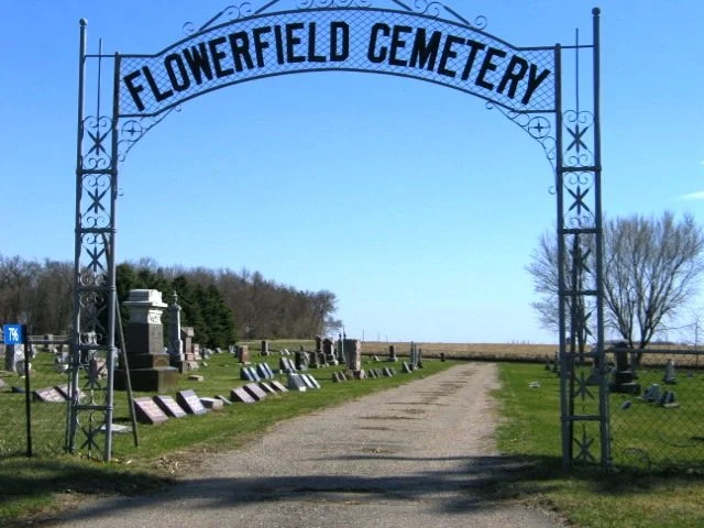 Entrance to Flowerfield Cemetery with a metal archway and a dirt path leading inside, surrounded by gravestones and trees in the background.