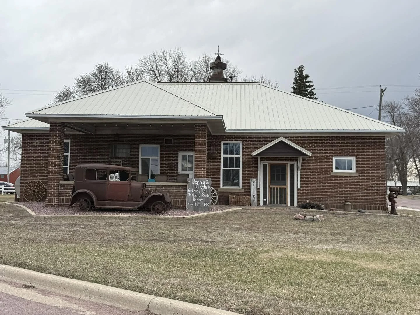 A historic brick building with a metal roof, featuring vintage car and a sign about Bonnie and Clyde's getaway car robbery in 1933, displayed outdoors.