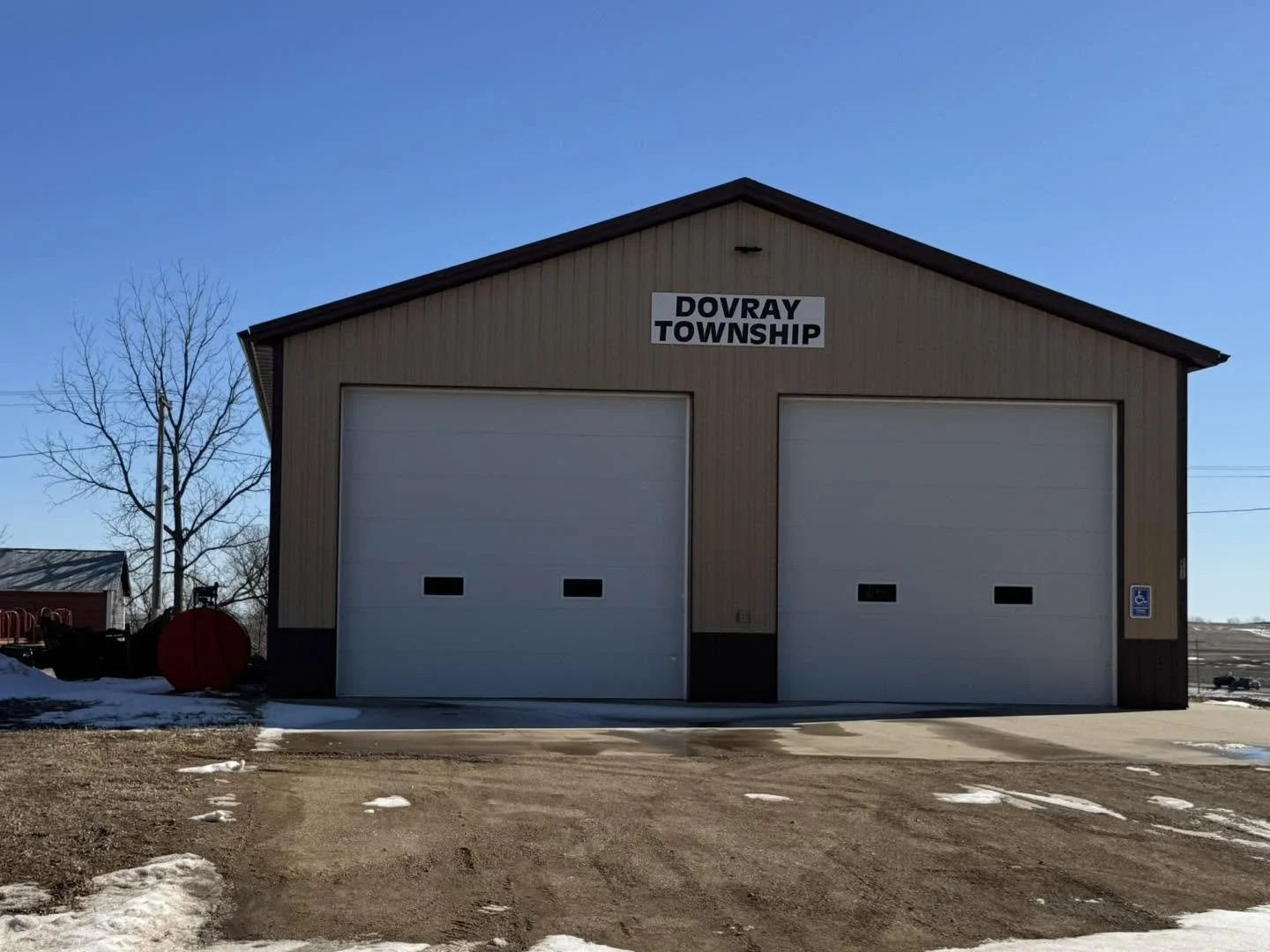A beige metal garage building with two white garage doors, situated in a rural area under a clear blue sky. A sign on the building reads 'DOVRAY TOWNSHIP.' There is a parking area with patches of snow and a handicapped parking sign on the right side.