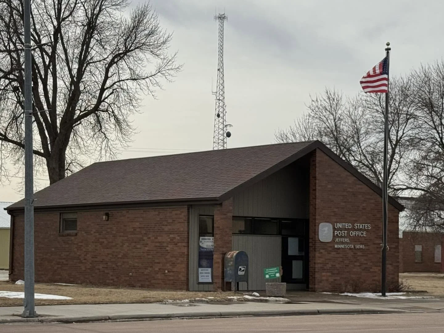 A brick United States Post Office building in Jefferson, Minnesota, with a flagpole displaying the American flag, and a postal mailbox in front.