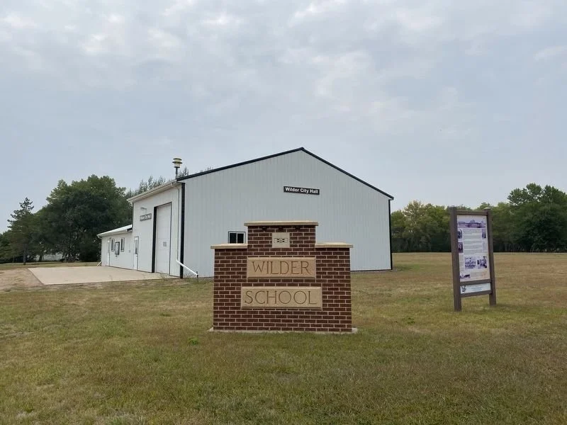A brick sign with the words 'Wilder School' in front of a large white building labeled 'Wilder City Hall' on a grassy field with trees in the background and an informational sign to the right.