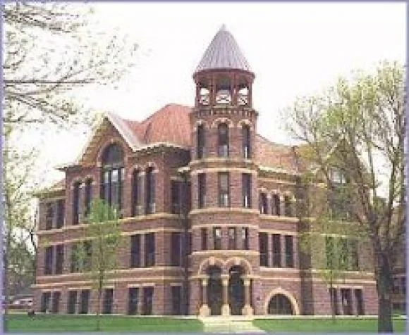 A historic brick building with a tower, arched windows, and a peaked roof, surrounded by trees.