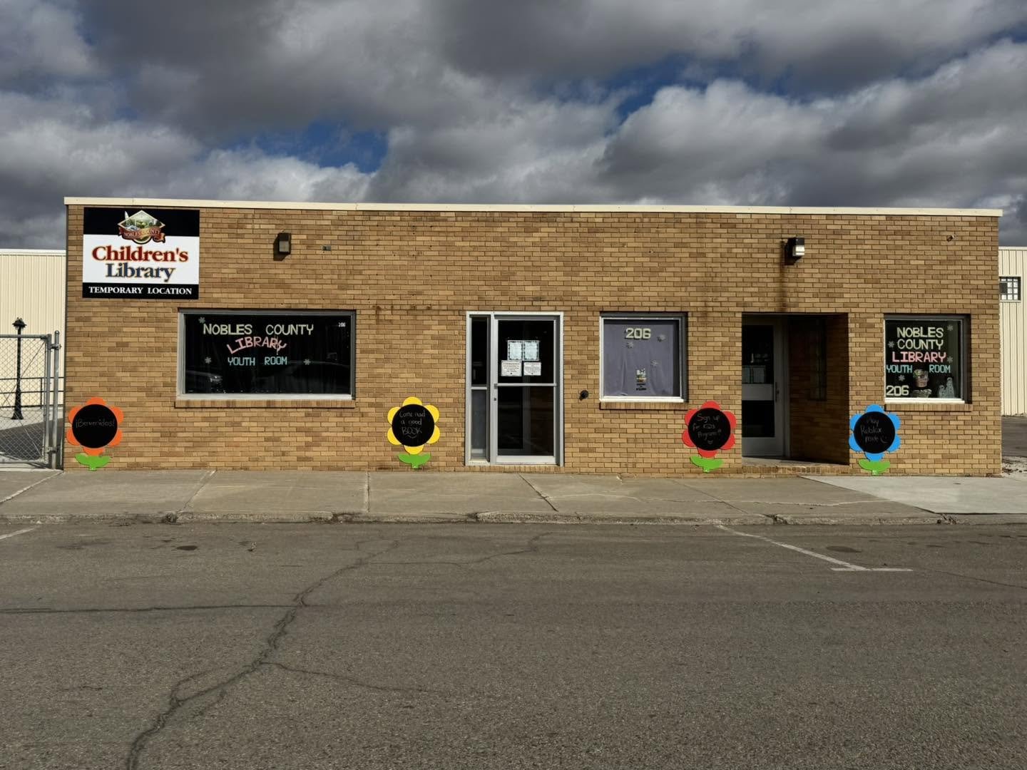 A brick building with signs indicating it is a children's library, specifically for Nobles County. There are four flower-shaped decorations on the sidewalk with handwritten messages. The sky is cloudy.