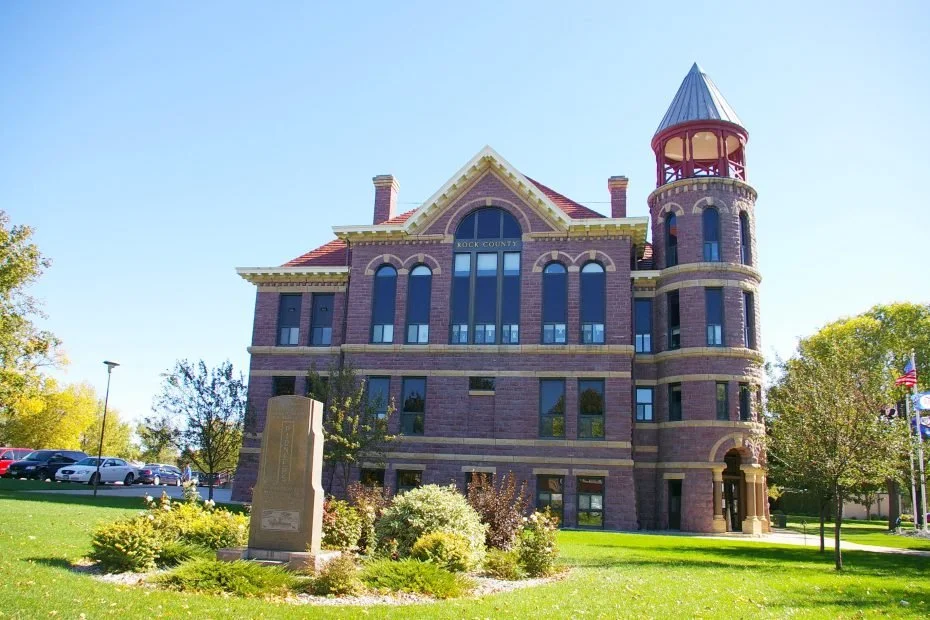 A historic red-brick courthouse with a turret, surrounded by a well-kept lawn and trees, with a parking lot and a monument in the foreground.
