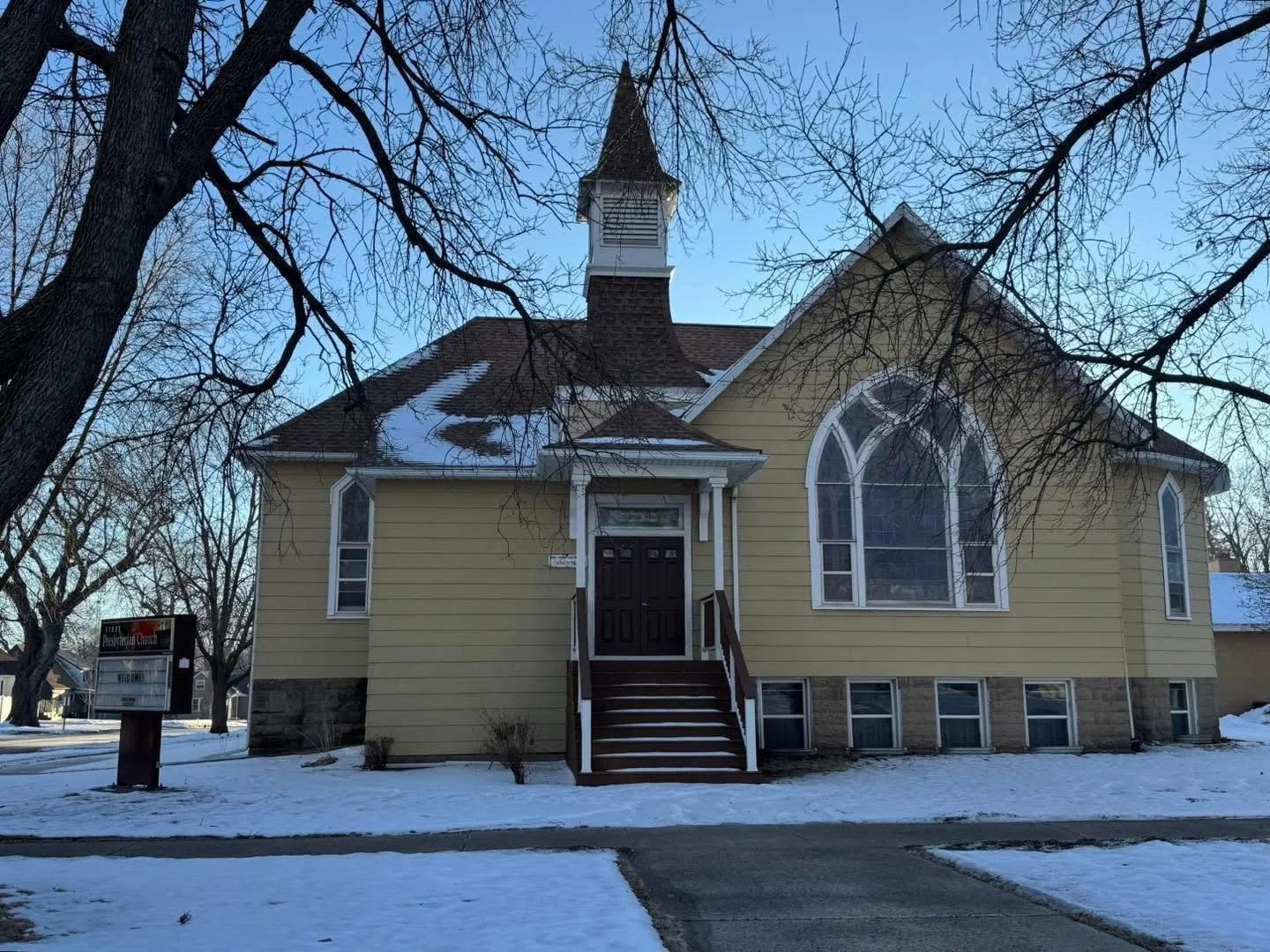 A yellow church with white trim, a steeple, and large arched stained glass windows, surrounded by snow on the ground and leafless trees.
