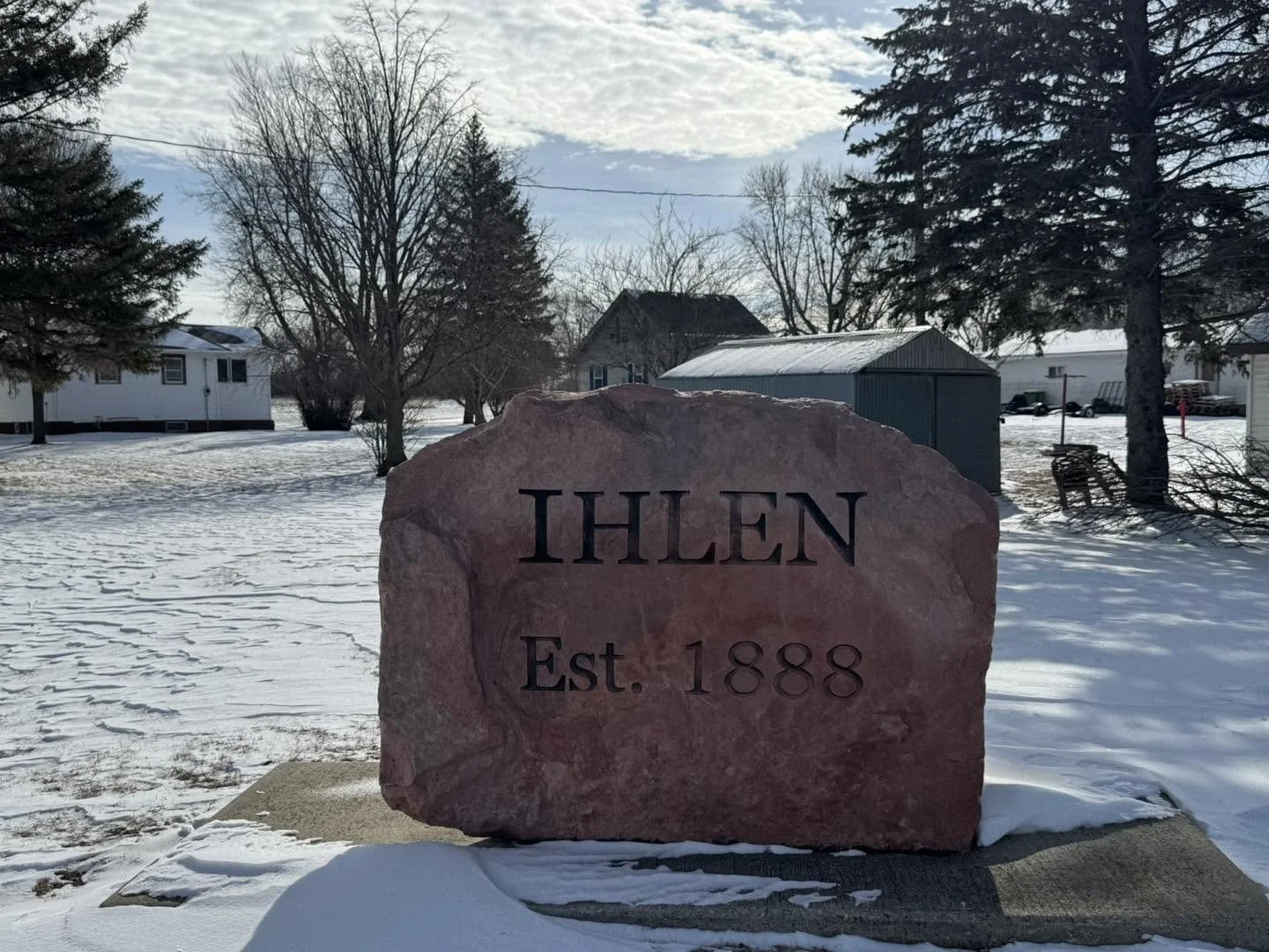 A large pinkish stone with the word IHLEN and the text Est. 1888 engraved on it, situated in a snow-covered yard with trees, houses, and a shed in the background.