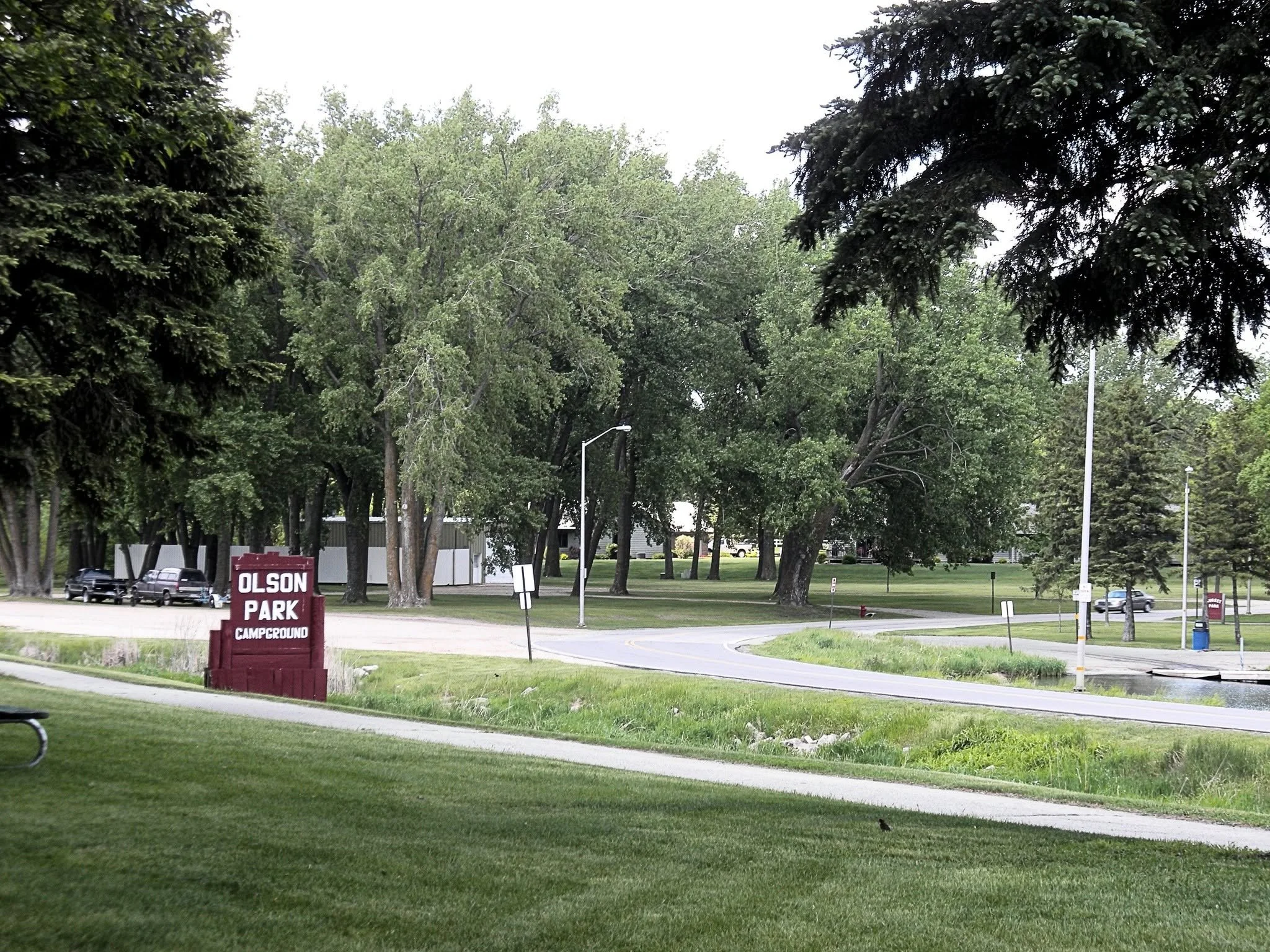 A park exit sign with the text 'Olson Park Campground' is visible, surrounded by green grass and trees, with cars parked and a parking lot in the background.