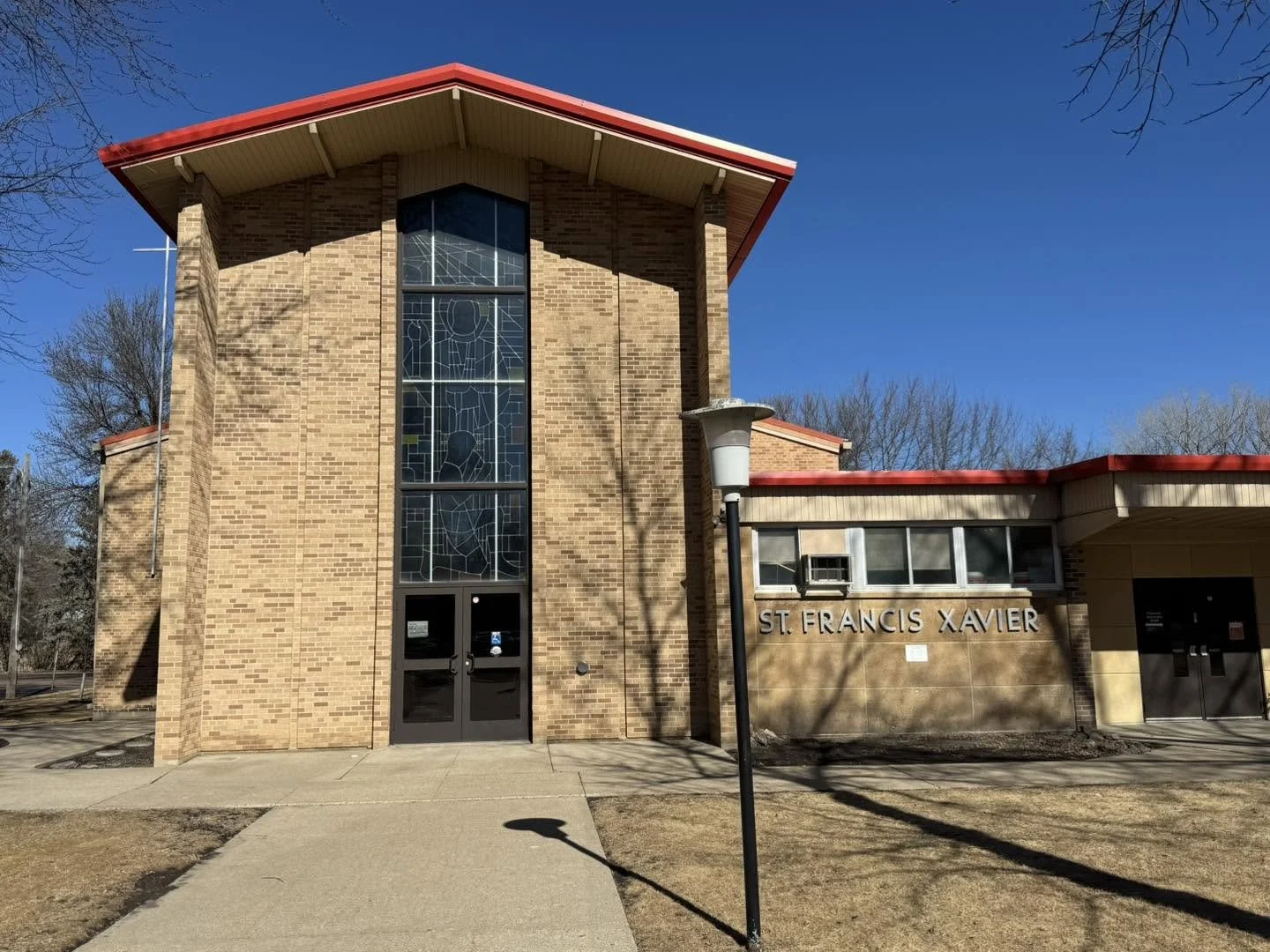 Exterior of St. Francis Xavier church building with brick facade, stained glass windows, and sign displaying the name, under a clear blue sky, with leafless trees and a sidewalk in front.
