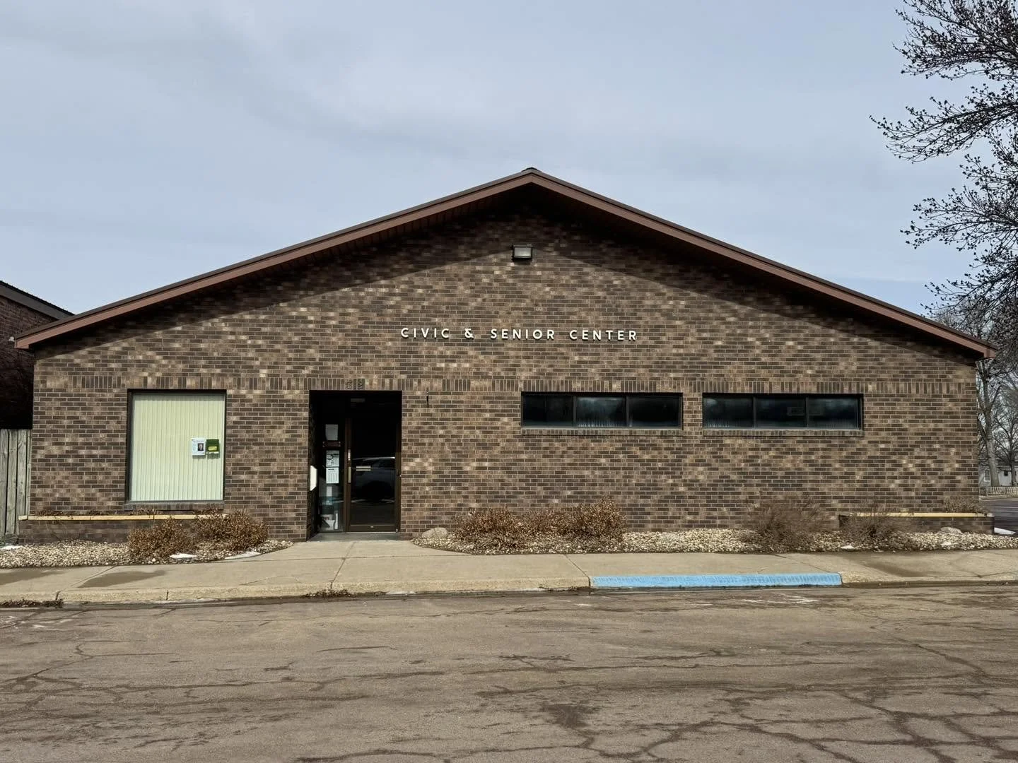 A brick building labeled 'Civic & Senior Center' with a front door, a large window with blinds, and a set of small windows, situated on a paved street with a parking space marked for handicapped parking.