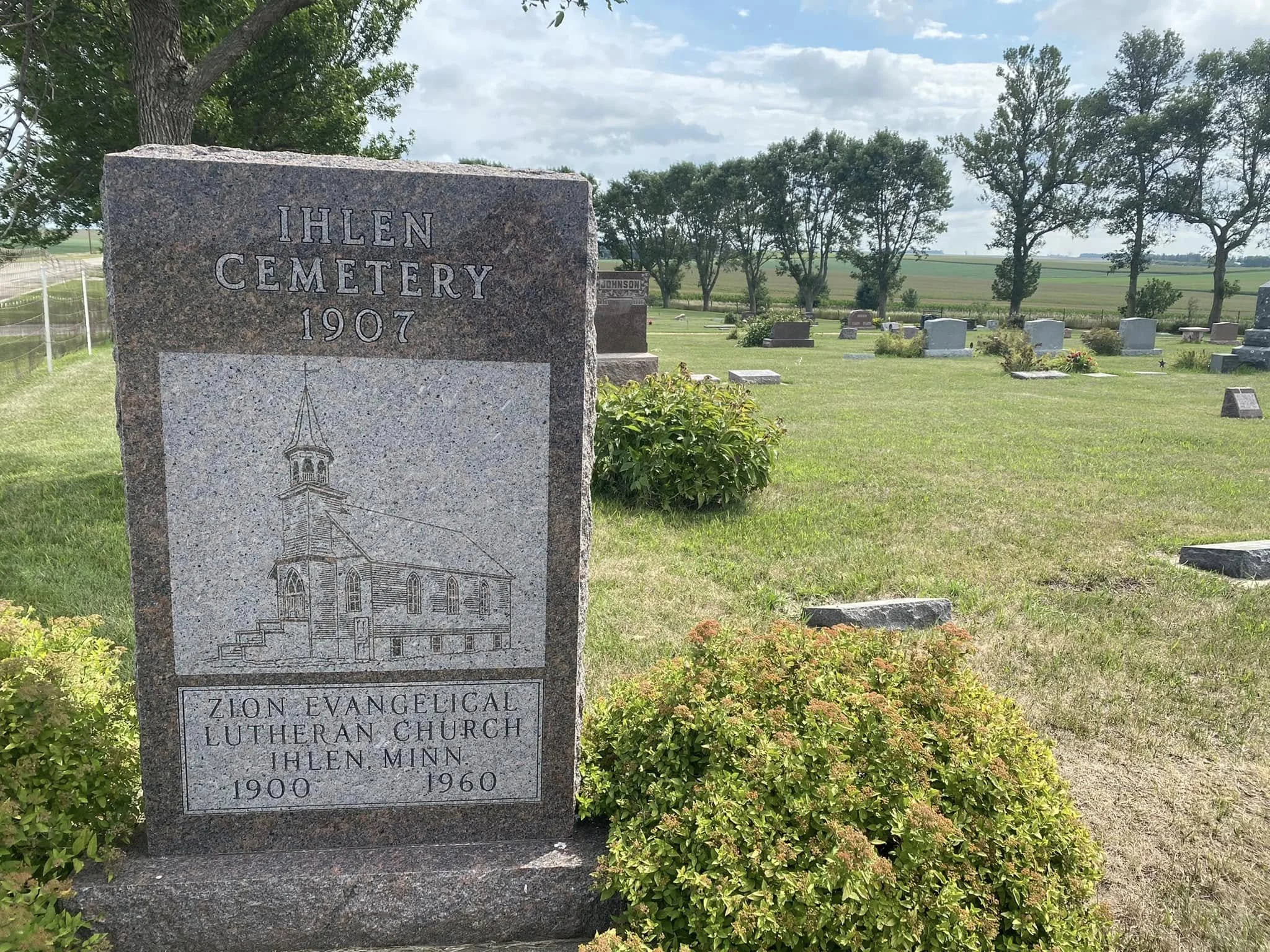 Gravestone at Ihlen Cemetery with the name ZION EVANGELICAL LUTHERAN CHURCH and dates 1900-1960, in a grassy area with other headstones and trees in the background under partly cloudy sky.