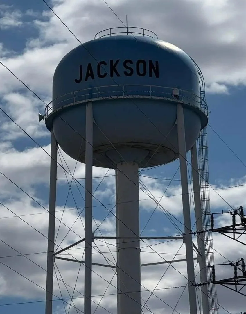 A large blue water tower with the word 'JACKSON' written on it, supported by tall white steel legs, against a partly cloudy sky.
