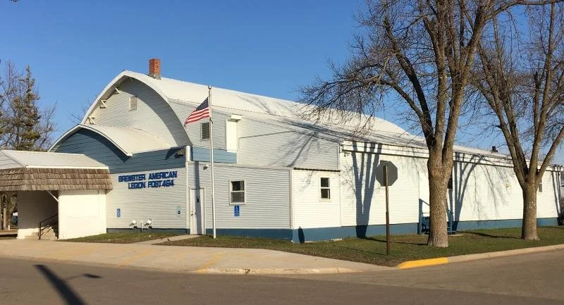 A white building with a curved roof, American flags, and a sign indicating it is a post for the American Legion, Post 464. There are trees and a street with a stop sign in front.