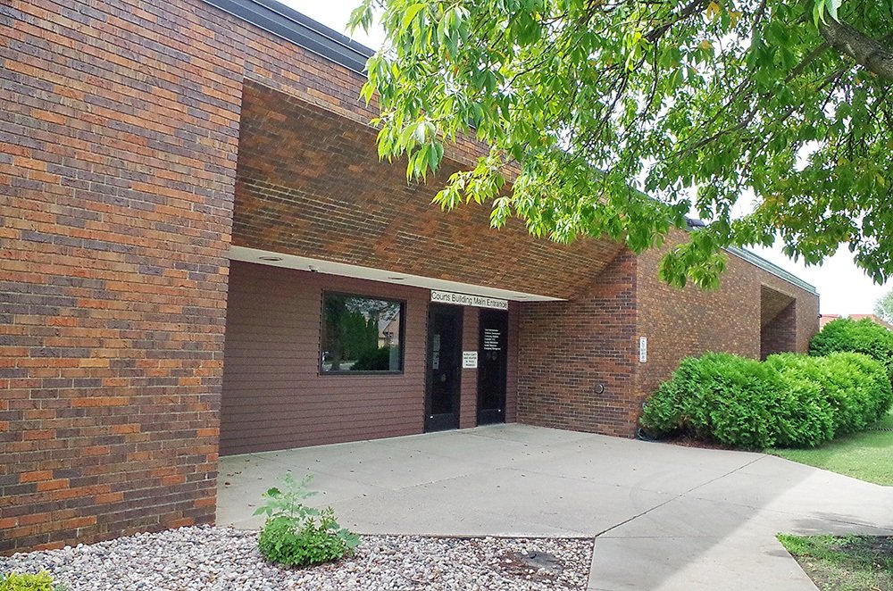 Exterior view of a brick courthouse building entrance with a sign indicating 'Courts Building Main Entrance', surrounded by green bushes and trees.