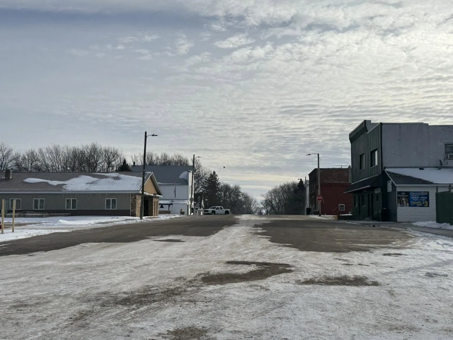 A quiet, snow-covered street in a small town, with single-story buildings on the left and two-story buildings on the right, under a partly cloudy sky.