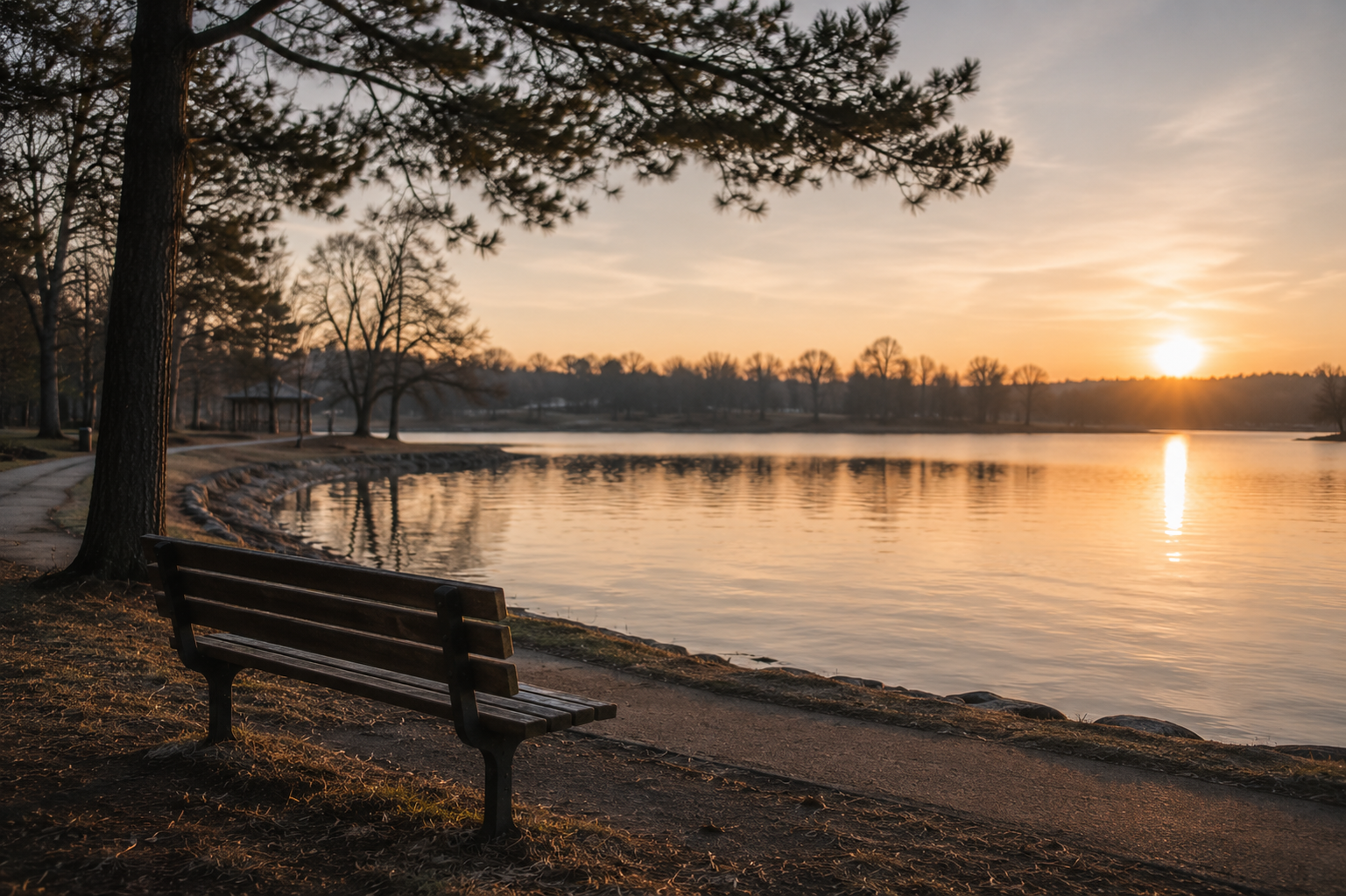 A peaceful lakeside scene at sunset with a park bench by the water and trees along the shore.