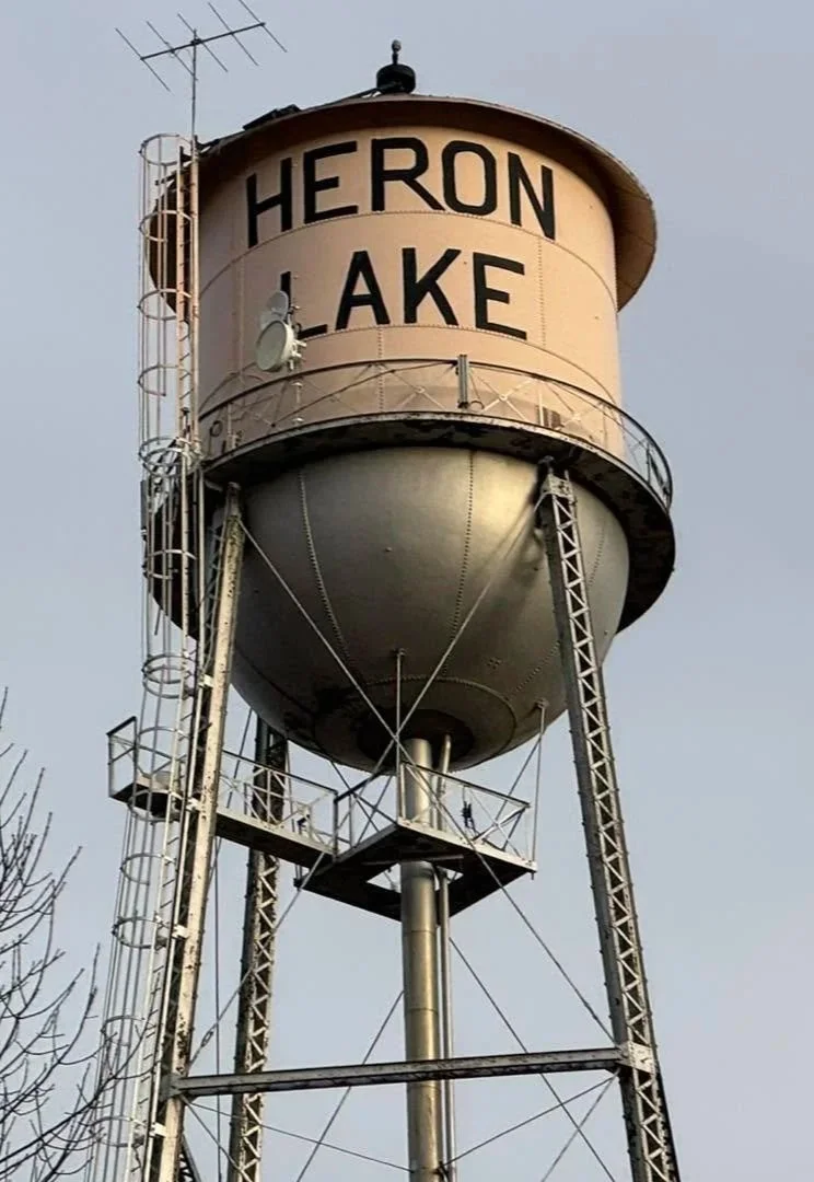 A water tower with the words 'Heron Lake' painted on it, supported by a metal framework with a small platform and ladder climbing it, and antennas on top.