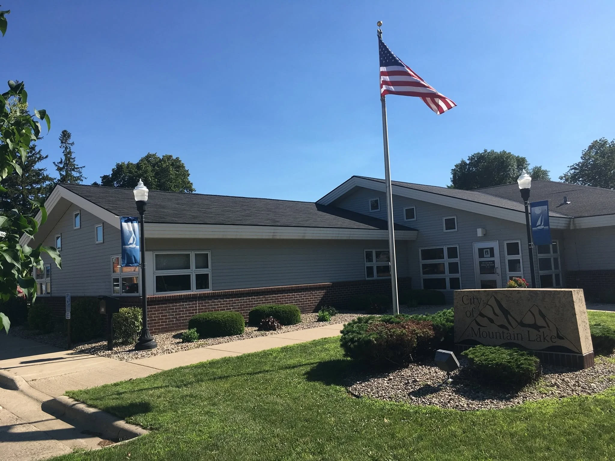 American flag flying on a flagpole in front of a light gray building with a front lawn and landscaping, under a clear blue sky.