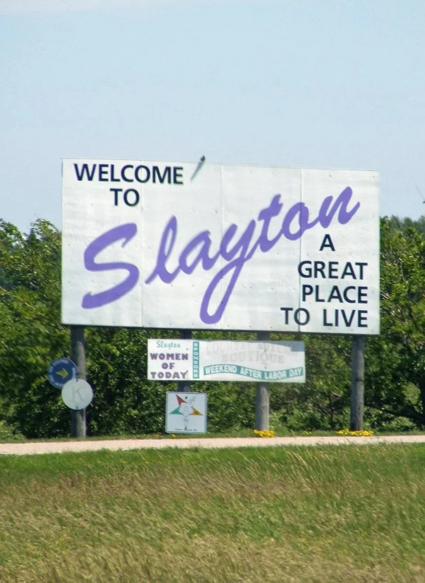 Large welcome sign for Slayton, describing it as a great place to live. Smaller signs below mention women of today, weekend after Labor Day, and a Texas-themed icon.