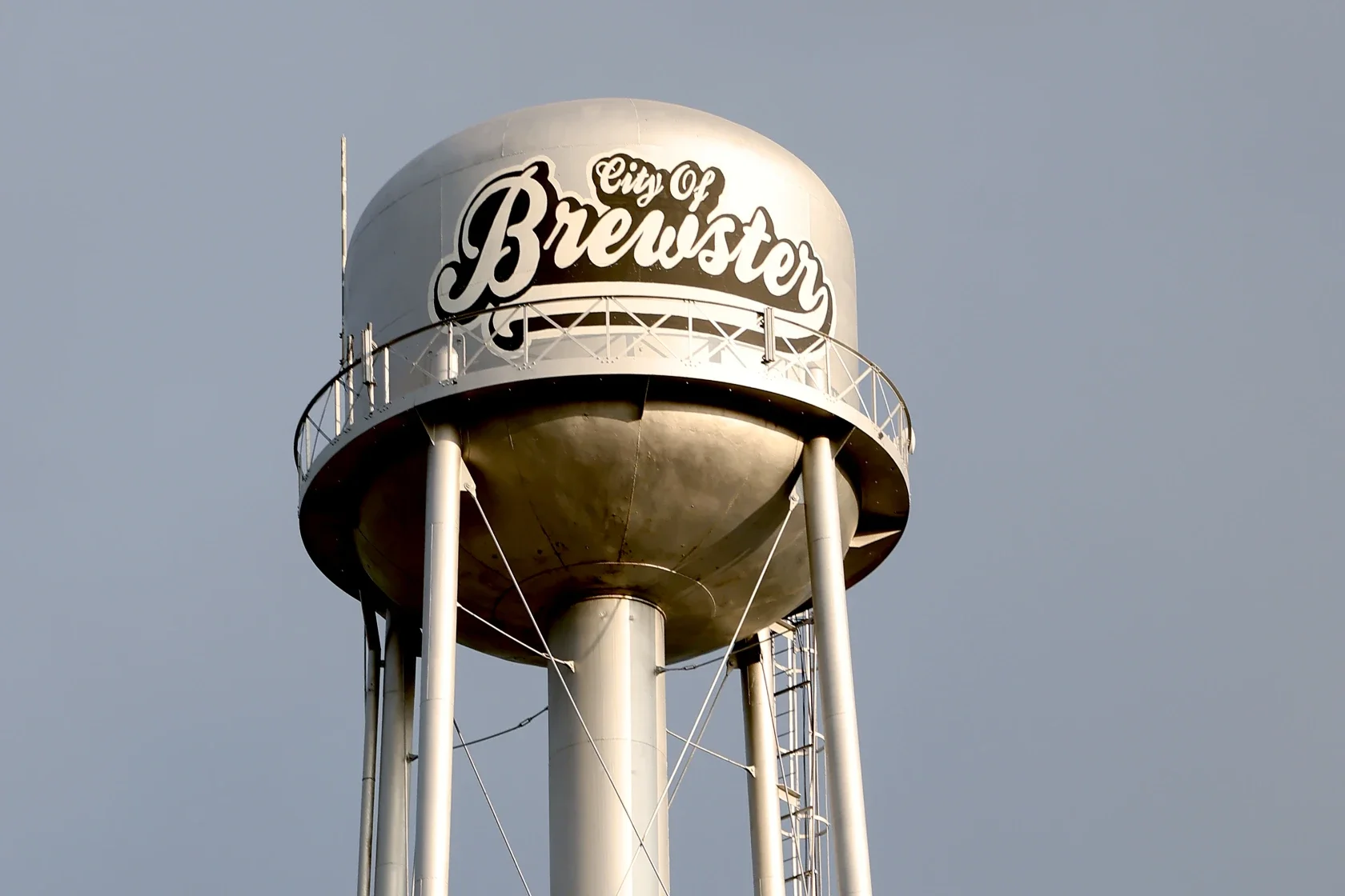 Water tower with the words 'City of Brewster' written on it, supported by four tall metal poles and a ladder for maintenance, against a gray sky.