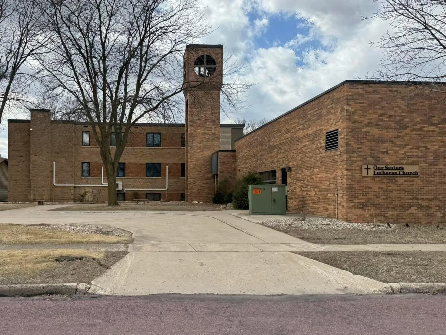 Red brick Lutheran church with a small sign that reads "Our Saviors Lutheran Church," a sidewalk, leafless trees, and a partly cloudy sky.