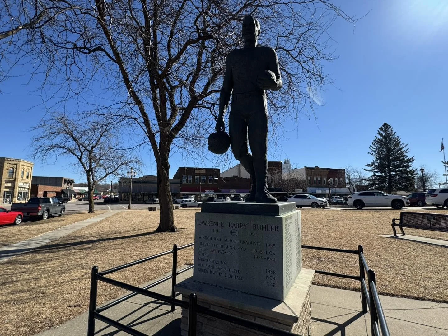 Statue of Lawrence Larry Buhler holding a ball, situated in a small park with leafless trees and a town street in the background on a clear day.