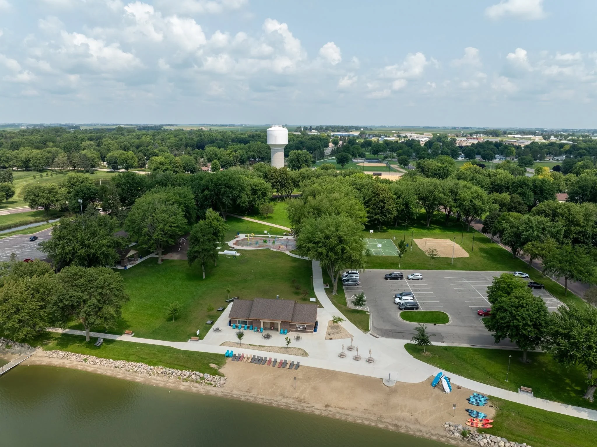 Aerial view of a park adjacent to a body of water with a sandy shoreline, kayaks, and a grassy area with trees, sports courts, and a water tower in the distance.