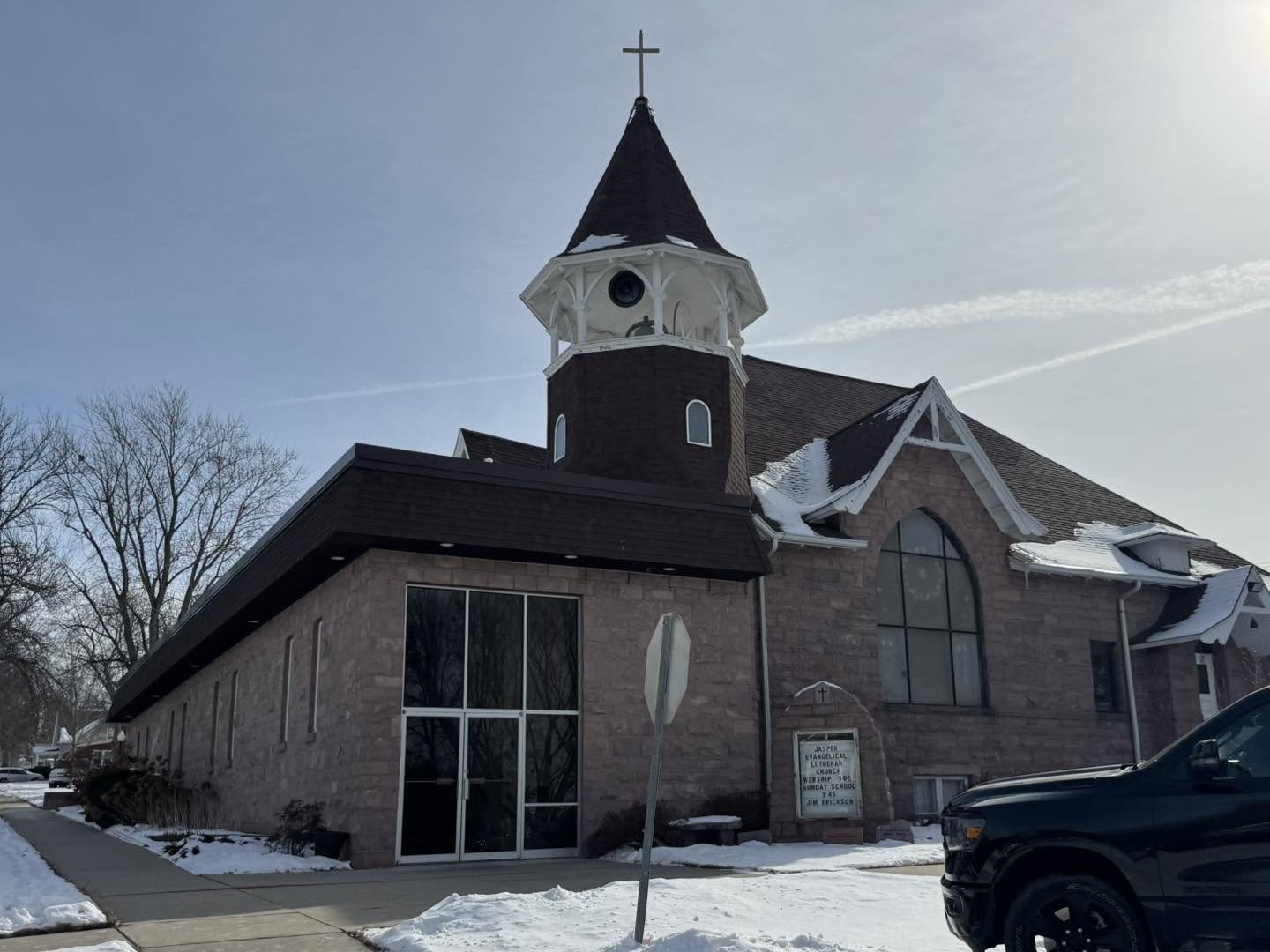 A church building with a stone facade, large arched windows, and a steeple topped with a cross, in a snowy landscape under a blue sky.