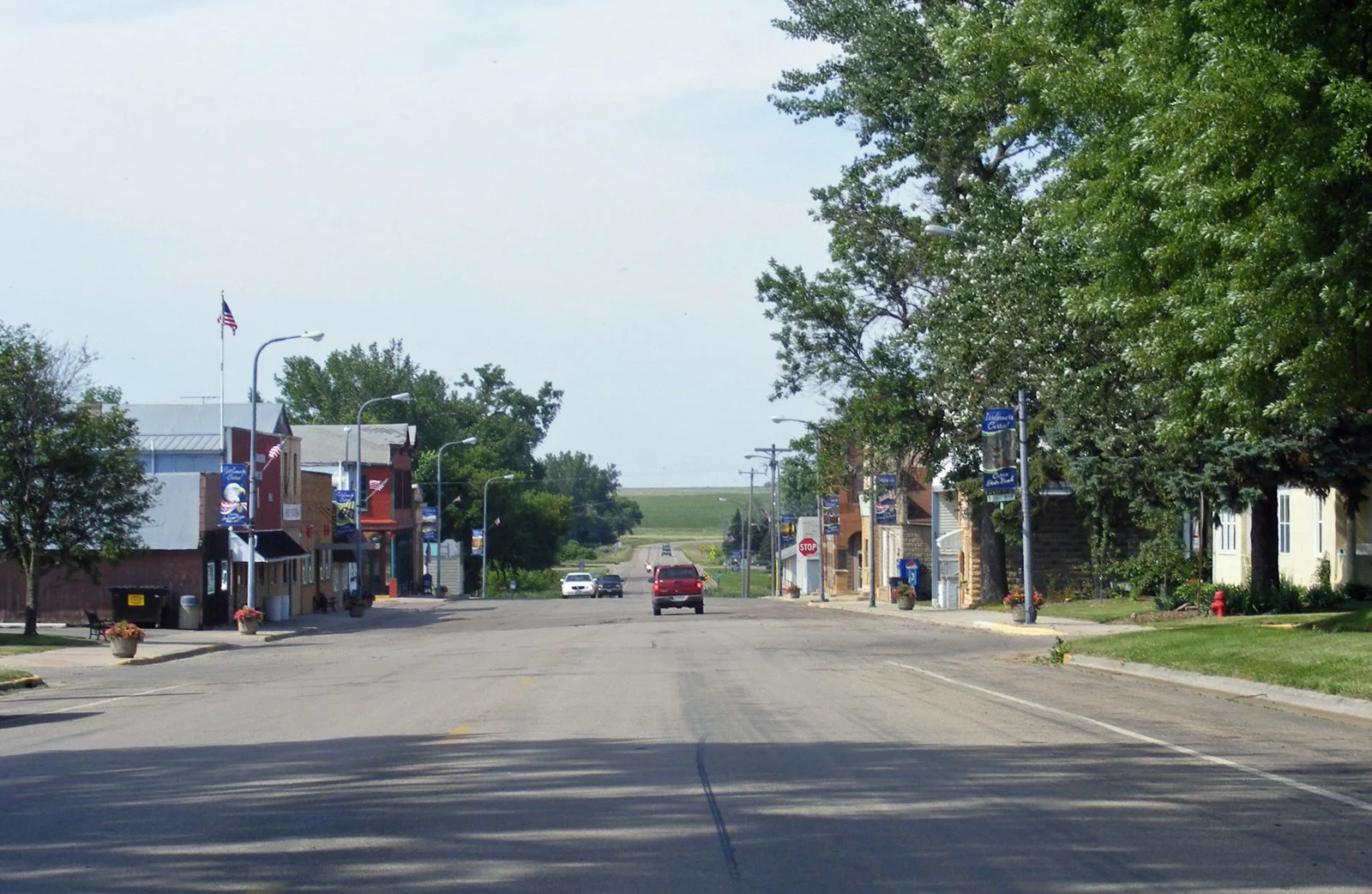 A small town street with colorful storefronts on both sides, trees, and a few cars driving through, with a stop sign visible in the distance.