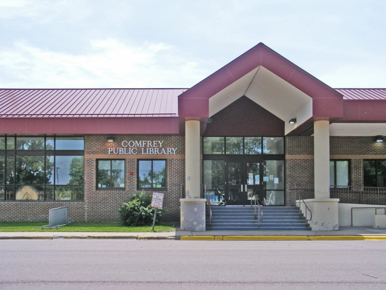 Exterior of Comfrey Public Library with brick and glass facade, steps leading to entrance, and a sign indicating no parking.