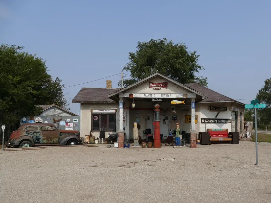 Old, rustic roadside store with vintage car, signs, and outdoor seating, located at corner of a small town.