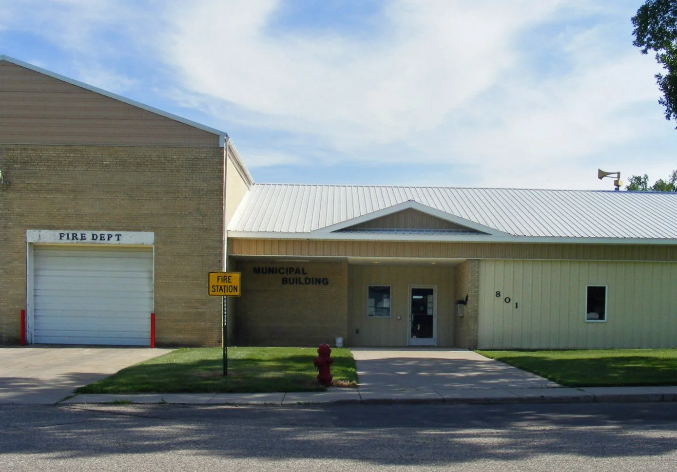 Image of a municipal building with a fire department garage on the left, a sign indicating fire station, and green lawn in front.
