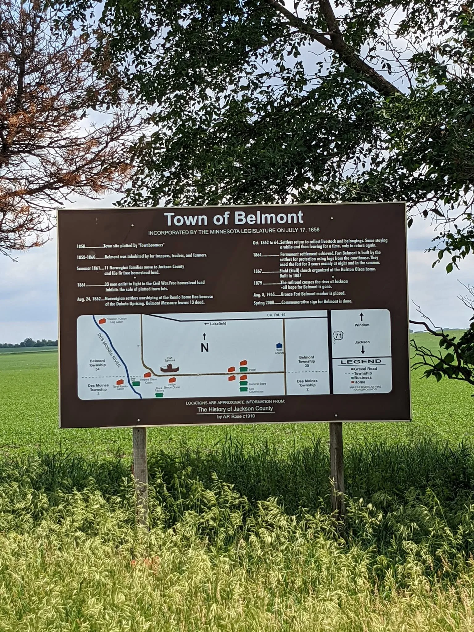 A historical informational signboard about the town of Belmont, Minnesota, with a map and historical timeline, surrounded by trees and green field.