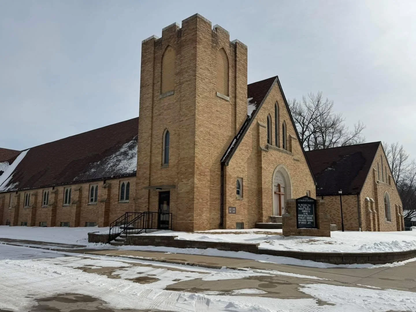 A brick church with a tall, square tower and a cross on the front door, surrounded by snow and leafless trees.