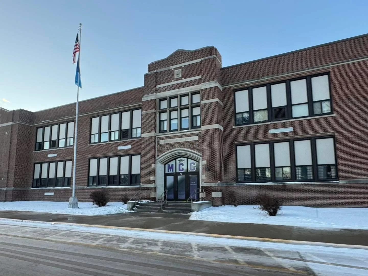 Brick school building with large windows and a central entrance marked with purple letters 'MCG'. A flagpole with the American and possibly state flags stands to the left of the building, with snow on the ground and a clear blue sky.