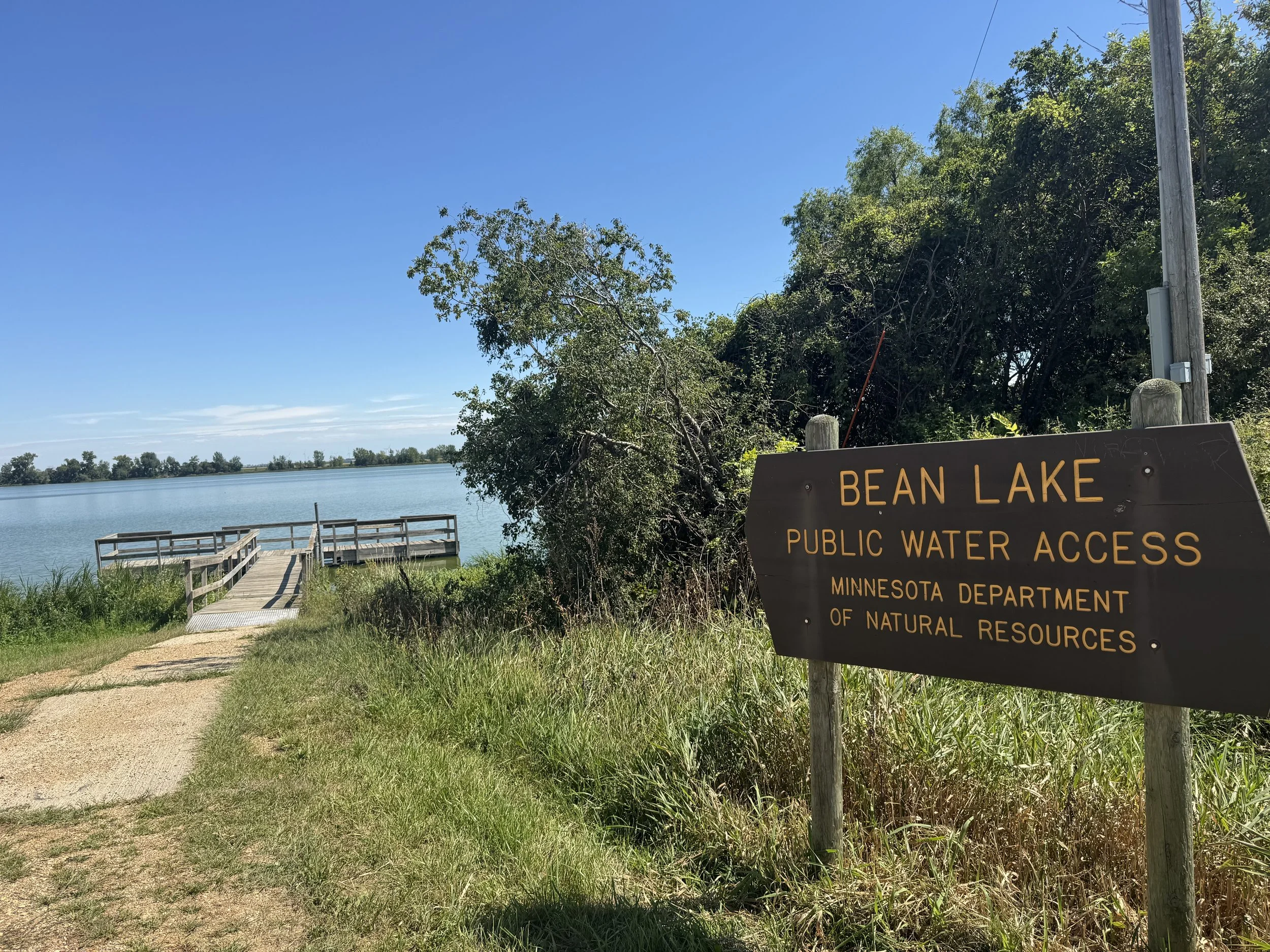 A view of Bean Lake with a wooden dock extending into the water, surrounded by trees and a clear blue sky. There is a sign indicating public water access from the Minnesota Department of Natural Resources.