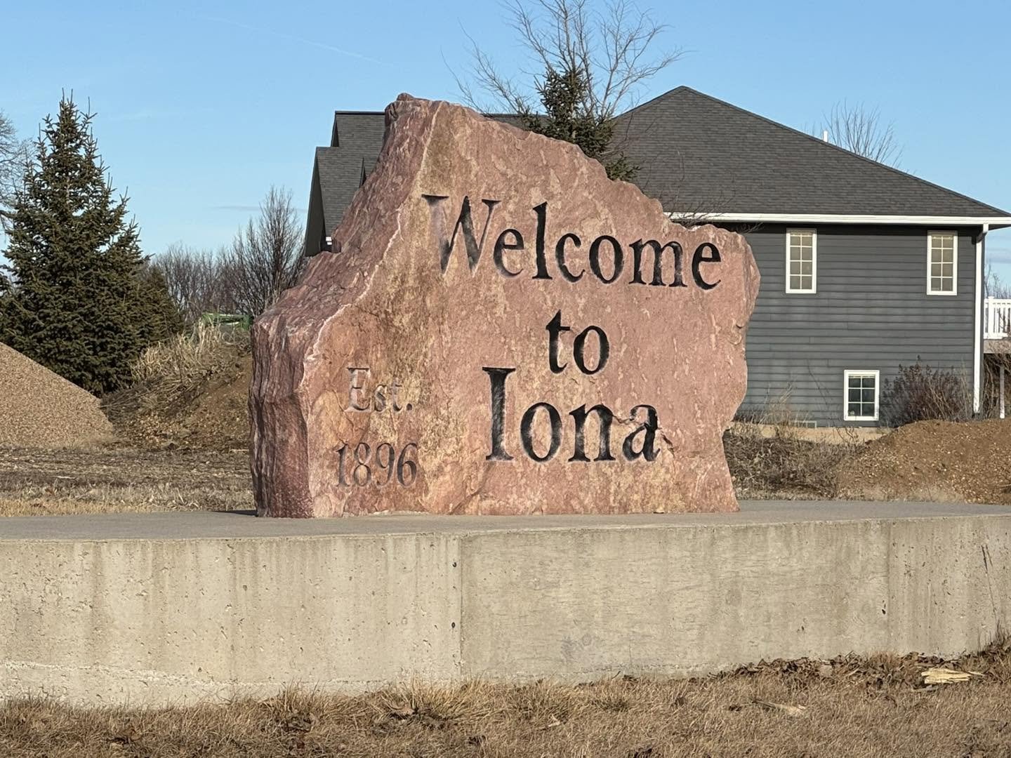 A large pinkish rock with the engraved words "Welcome to Iona," and "Est. 1896" on it, set on a concrete base near a residential neighborhood with trees and houses in the background.