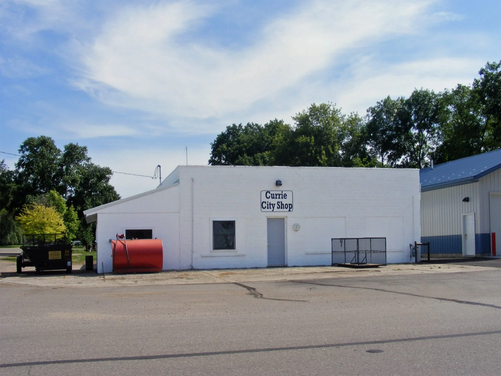 A white building with a sign that reads 'Currie City Shop' on the front, with a red propane tank and a black trailer parked outside. Green trees and a blue sky with clouds are in the background.