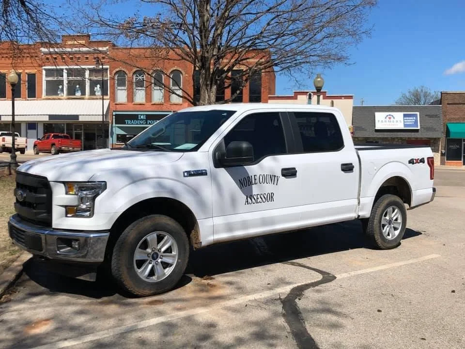 White pickup truck with 'Noble County Assessor' on the side, parked on the street in front of shops, with a leafless tree and a blue sky in the background.